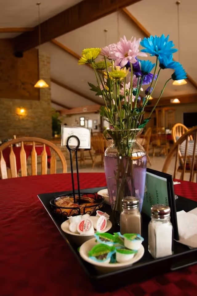 A dining table with a red tablecloth in a senior living facility dining room. On the table is a black tray holding a clear glass vase with colorful flowers, small plates with creamers and butter packets, salt and pepper shakers, and a menu holder. Wooden chairs and warm lighting fixtures are visible in the background.