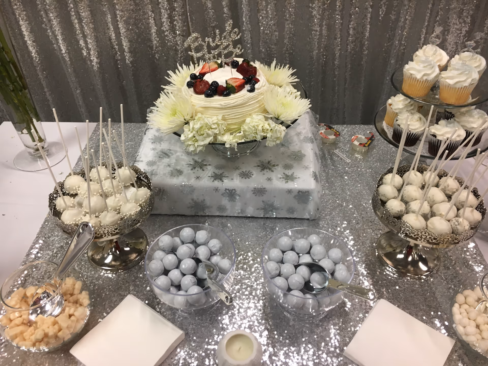 A sparkling dessert table with cake, cupcakes, and candies against a silver glitter backdrop.