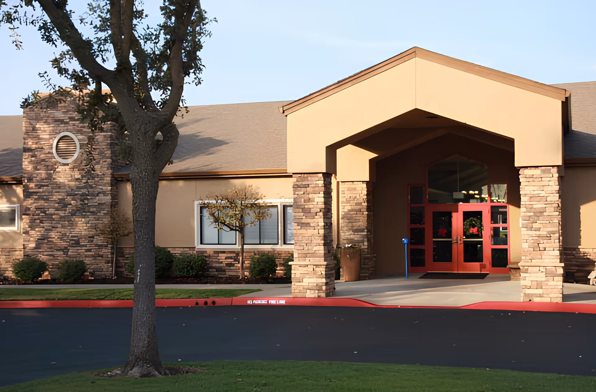 Front entrance of a senior living facility with a covered stone-pillared portico, red double doors, and a tree in the foreground.