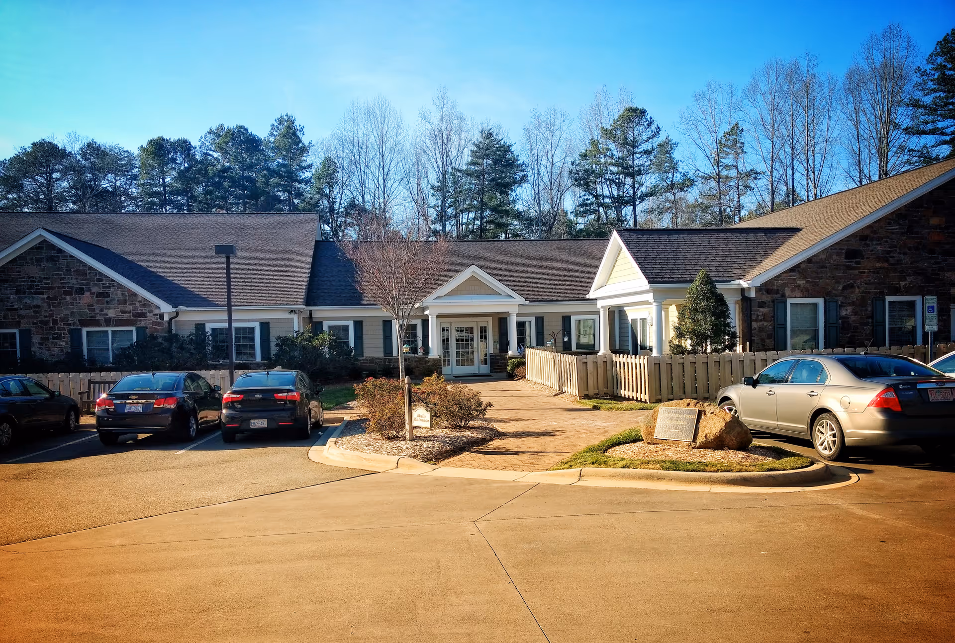 Front view of a hospice building with a parking lot, entrance, and surrounding trees.