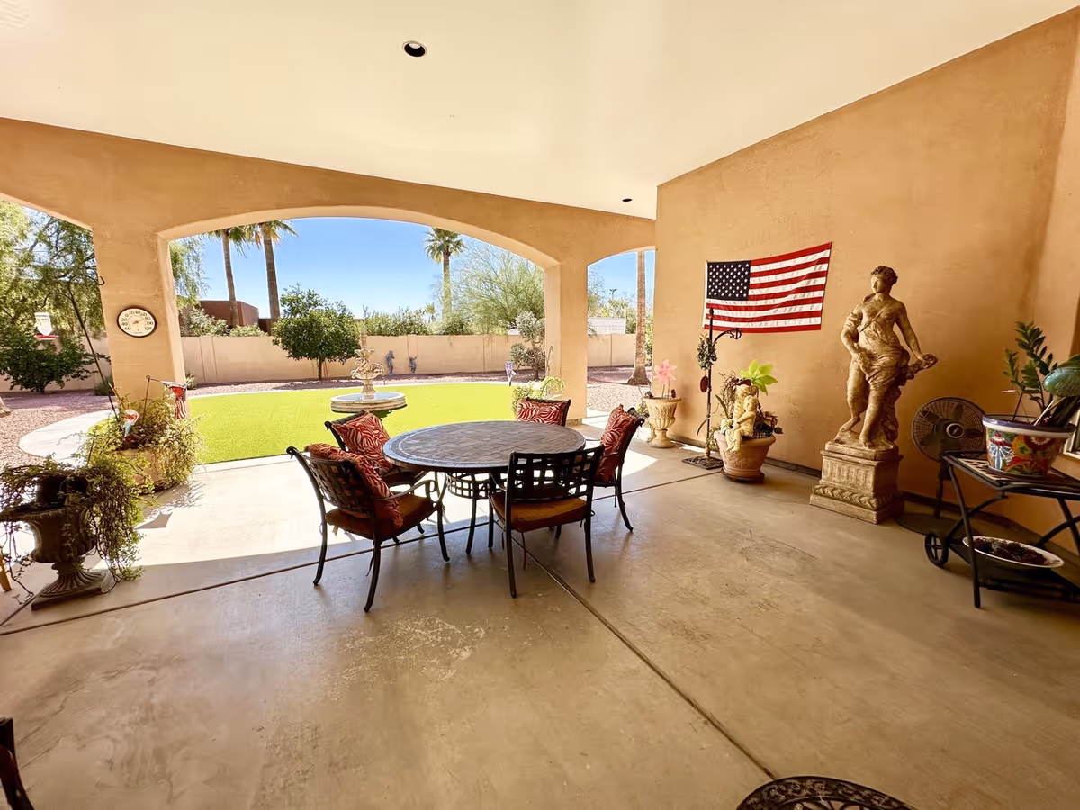 Covered patio with round table, chairs, potted plants, statue, flag, and grassy yard beyond.