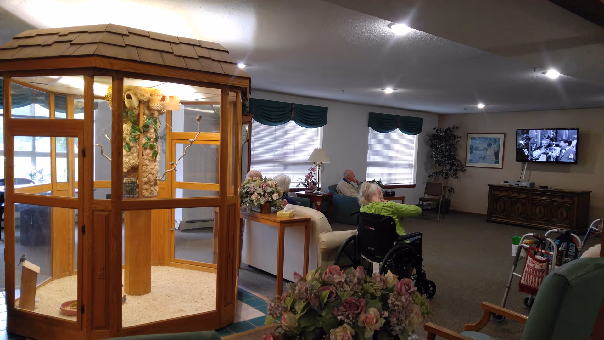 Senior living common room with a wooden framed glass enclosure and residents seated watching TV.