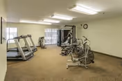 A fitness room with exercise equipment including two treadmills, a stationary bike, an elliptical machine, and a multi-functional weight machine. The room has beige carpet, white walls, fluorescent ceiling lights, a clock on the wall, and a window with vertical blinds.