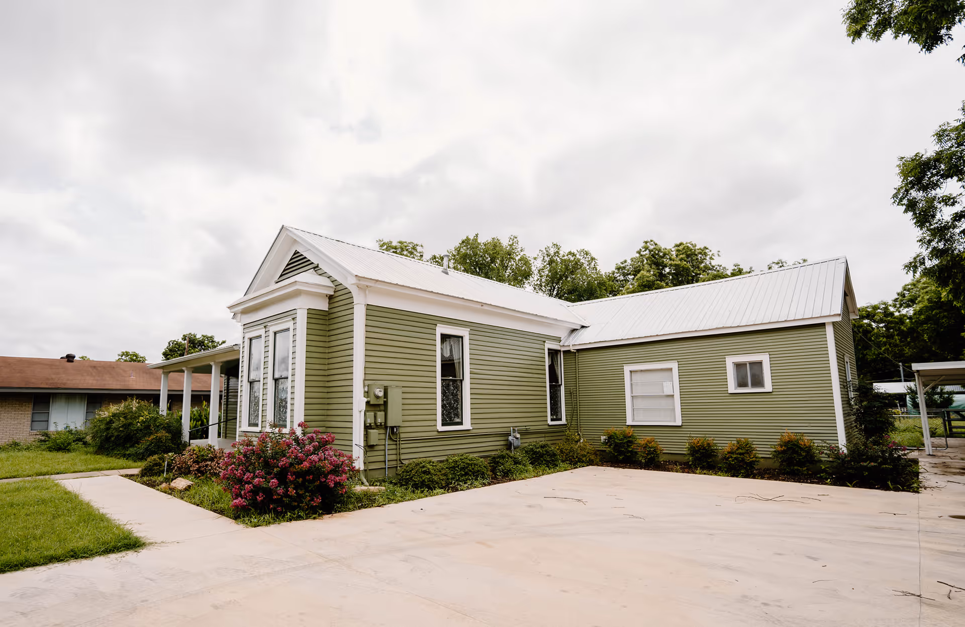 A single-story green building with white trim and a metal roof, surrounded by shrubs.