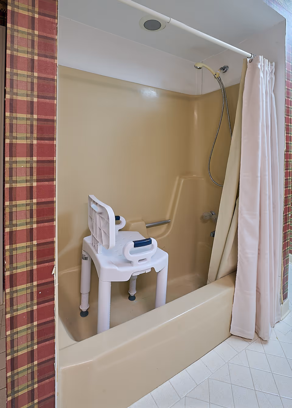 Beige shower stall with built-in seat and portable bath chair, surrounded by plaid walls.