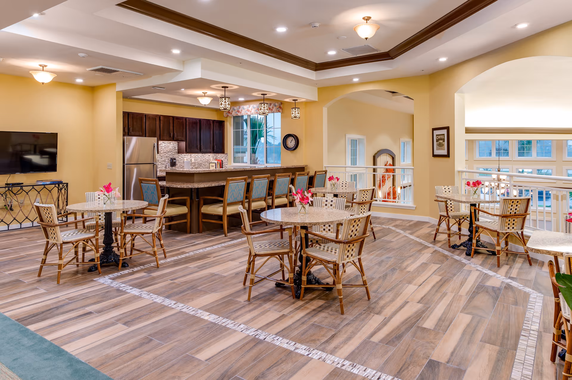 A spacious dining area with several round tables and wicker chairs, each table decorated with a small vase of pink flowers. The room features a kitchen area with a bar counter and stools, dark wood cabinets, stainless steel refrigerator, and pendant lighting. The floor has a wood-like tile design, and the walls are painted a warm yellow. Large windows and an open railing overlook another area below.