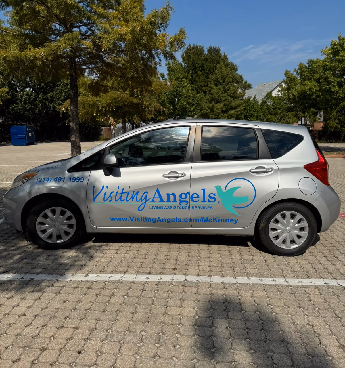 A silver Visiting Angels car parked in a lot with trees in background