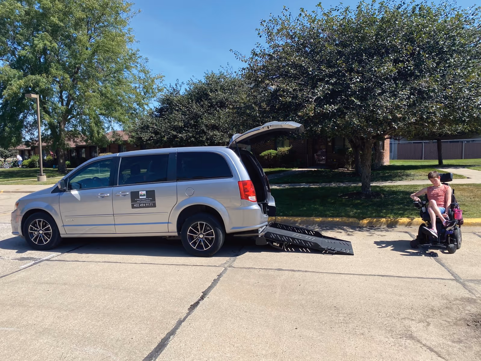 Silver wheelchair-accessible van with ramp parked in lot beside a person in a motorized wheelchair.