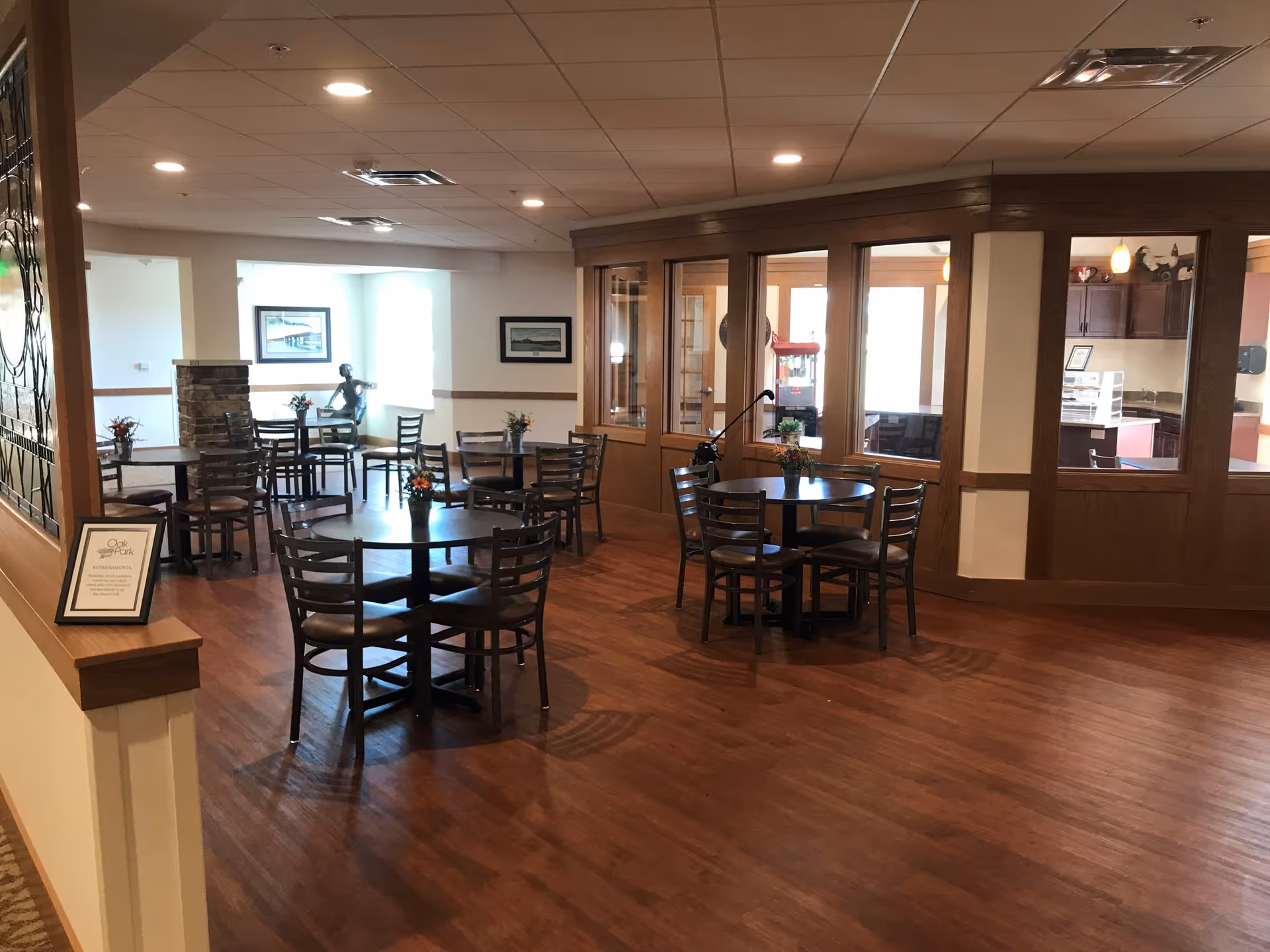 A dining area in Oak Park Place Green Bay with several round tables and chairs arranged on a wooden floor. The room has large windows and a glass-enclosed kitchen or serving area in the background. There are small flower arrangements on the tables and framed pictures on the walls.