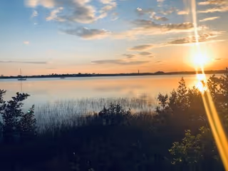 Sunset over a calm body of water with some bushes and tall grass in the foreground and a partly cloudy sky.