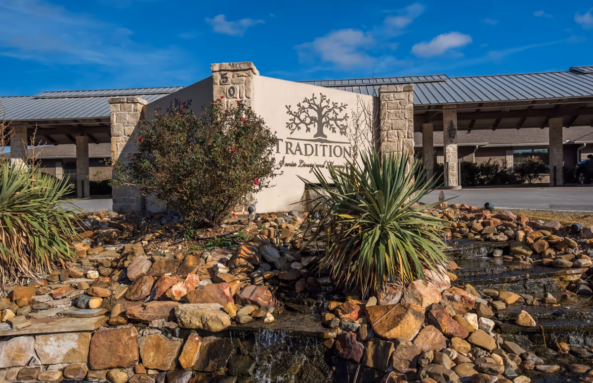 Stone entrance sign for Traditions Senior Living with a tree logo, surrounded by rocks, plants and a small cascading water feature in front of the facility.