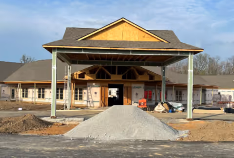 Front view of a building under construction with a large covered entrance supported by four pillars. The building has a pitched roof and multiple windows, with construction materials and equipment visible around the site.