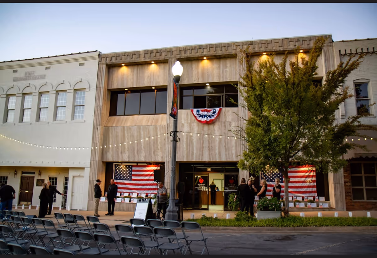 Front exterior of a beige building with American flags, string lights, and chairs outside.
