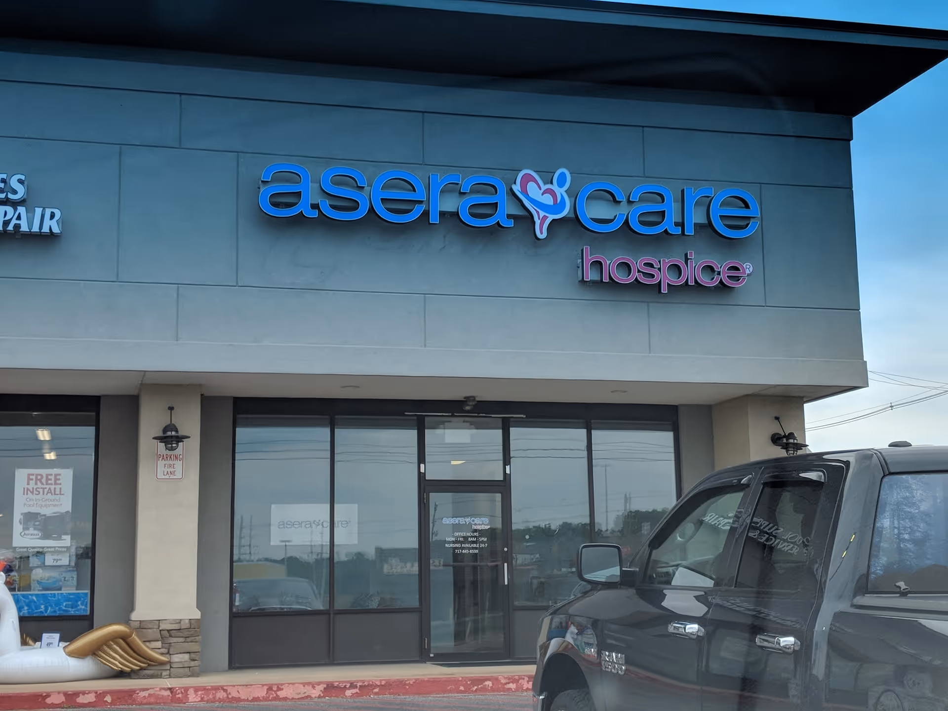 Exterior of a hospice storefront with blue AseraCare sign and a parked black SUV.
