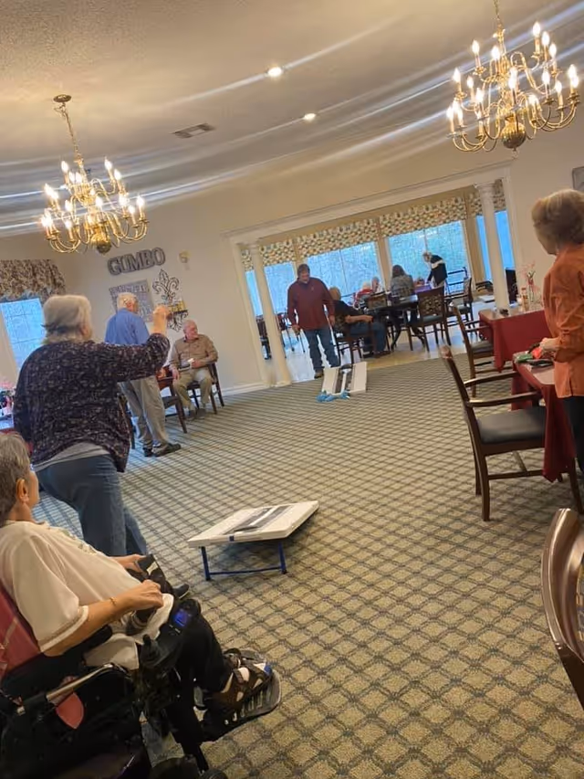 Seniors gathered in a carpeted dining/activity room with chandeliers, playing a cornhole-style toss game while others sit at tables.