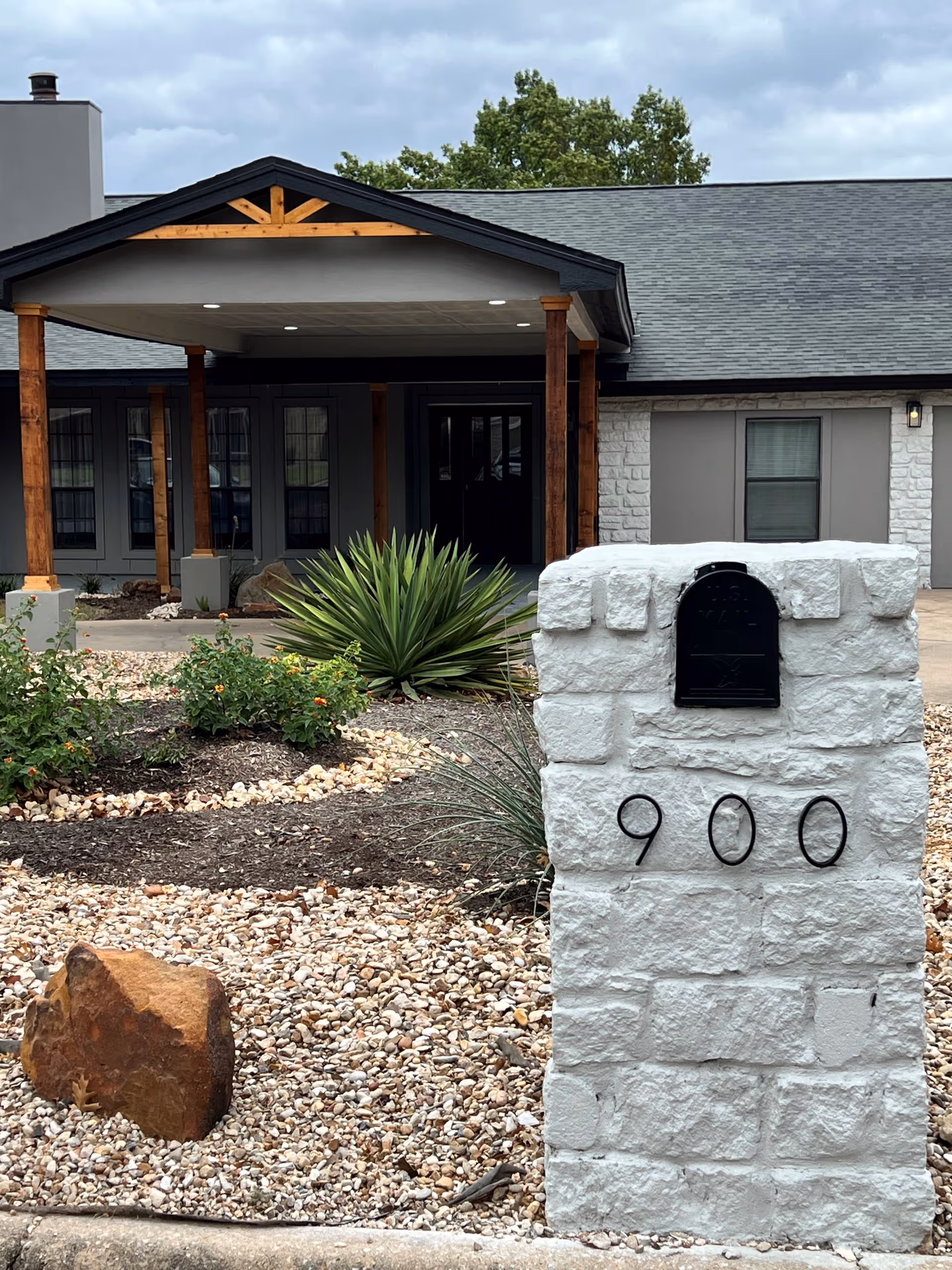 Front exterior view of a single-story building with a covered entrance supported by wooden pillars. In the foreground, there is a white stone mailbox with the number 900 and a black mailbox attached. The landscaping includes rocks, a large rock, and various plants.