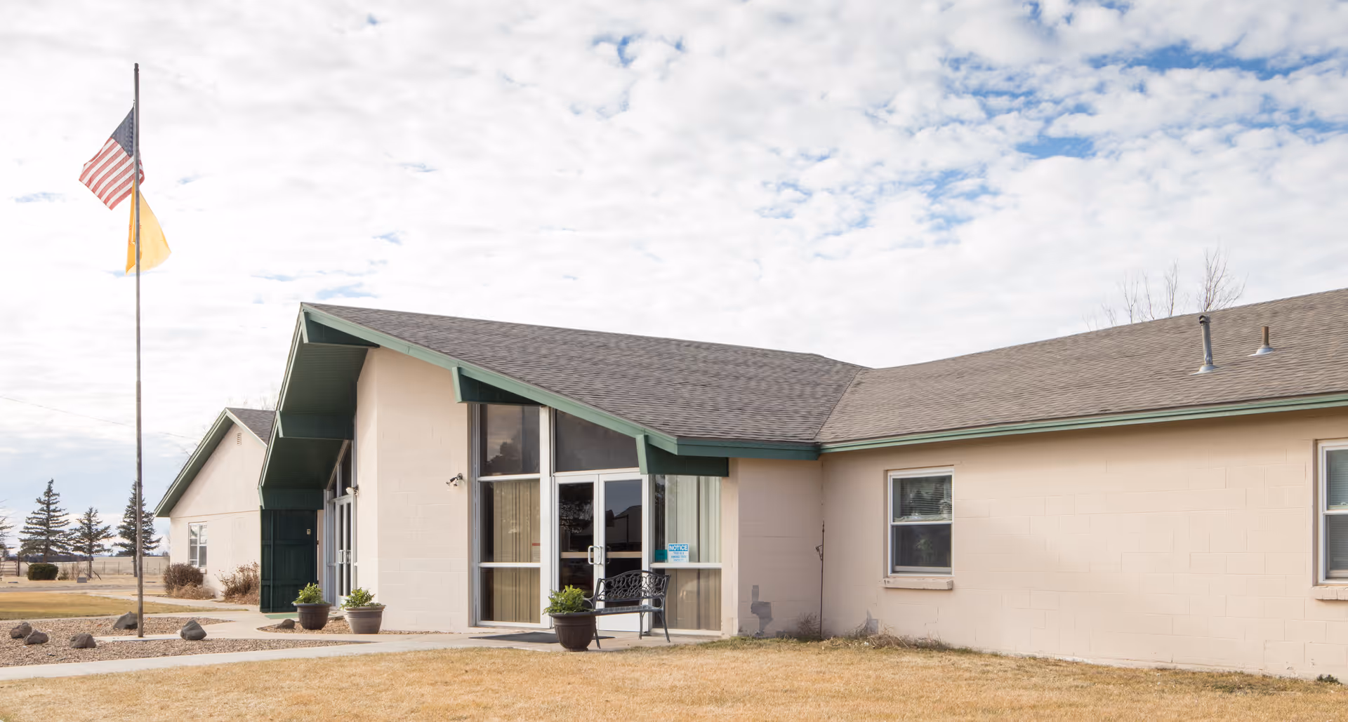 Exterior view of Clayton Nursing & Rehab facility showing a single-story building with beige walls and a green roof trim. There is a glass entrance door with potted plants and a bench outside. An American flag and another flag are flying on a flagpole near the building. The sky is partly cloudy.