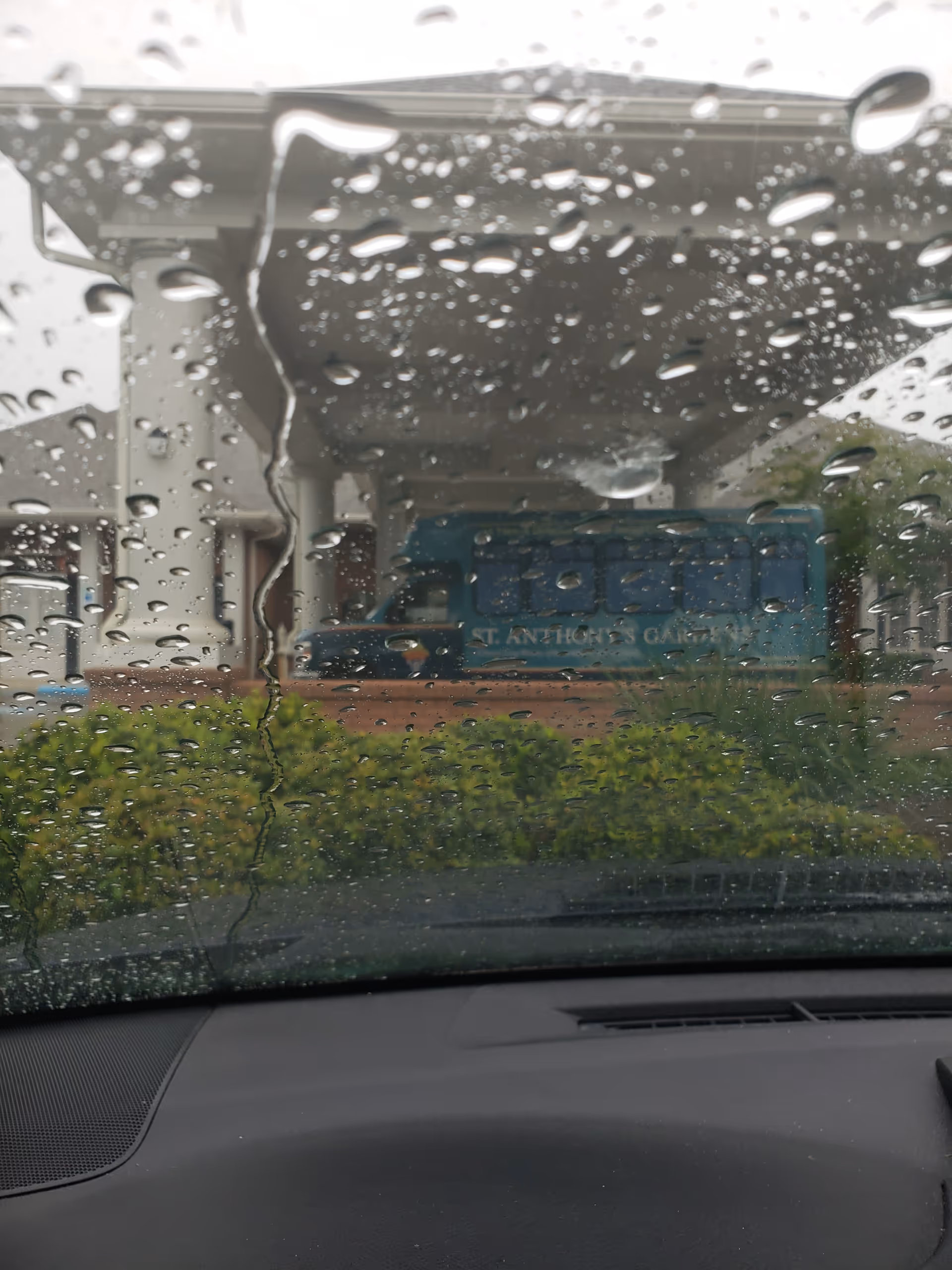 Rainy windshield view of St. Anthony's Gardens entrance with a blue bus under a porte-cochère