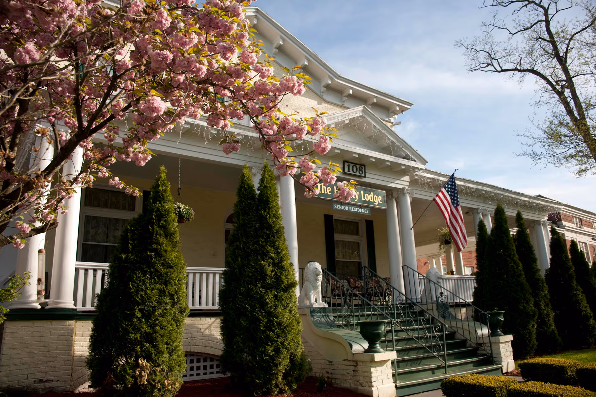 Front view of Ivy Lodge Assisted Living building with steps, columns, and blooming tree.