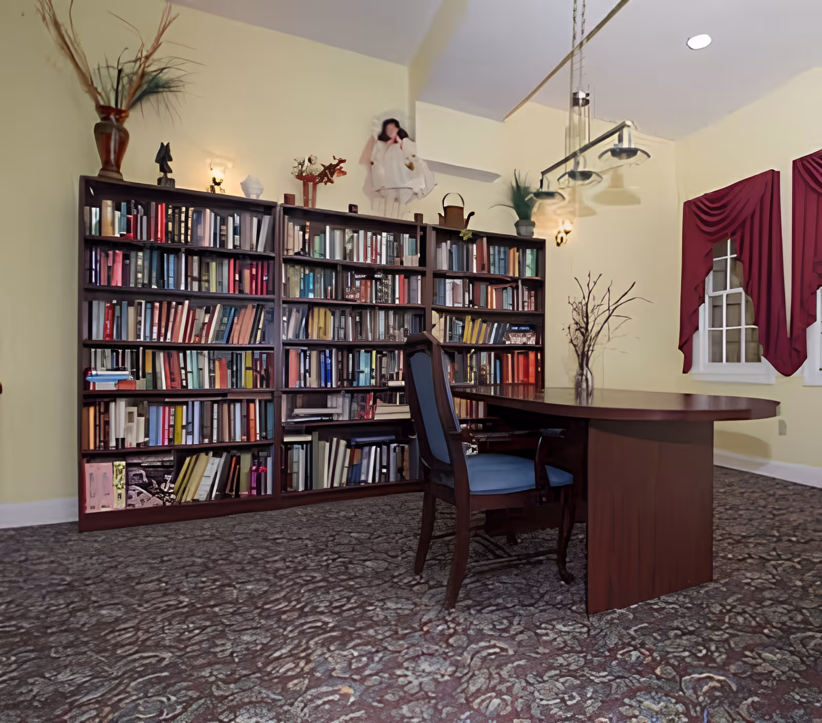 A cozy library-style common room with a large bookshelf, a wooden table and chair, hanging lights, and windows with red drapes.
