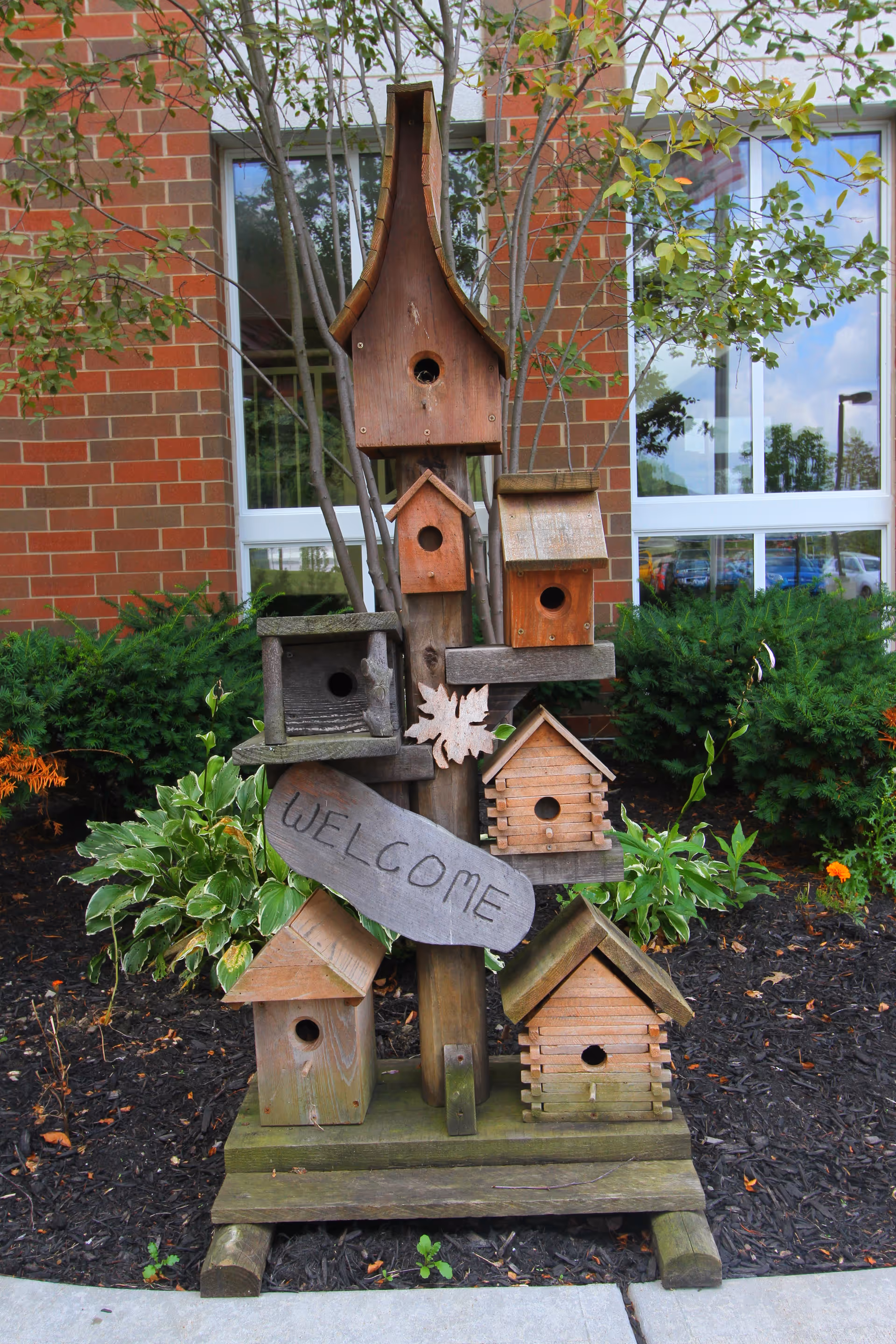 Tall wooden birdhouse sculpture with stacked birdhouses outside a brick building.