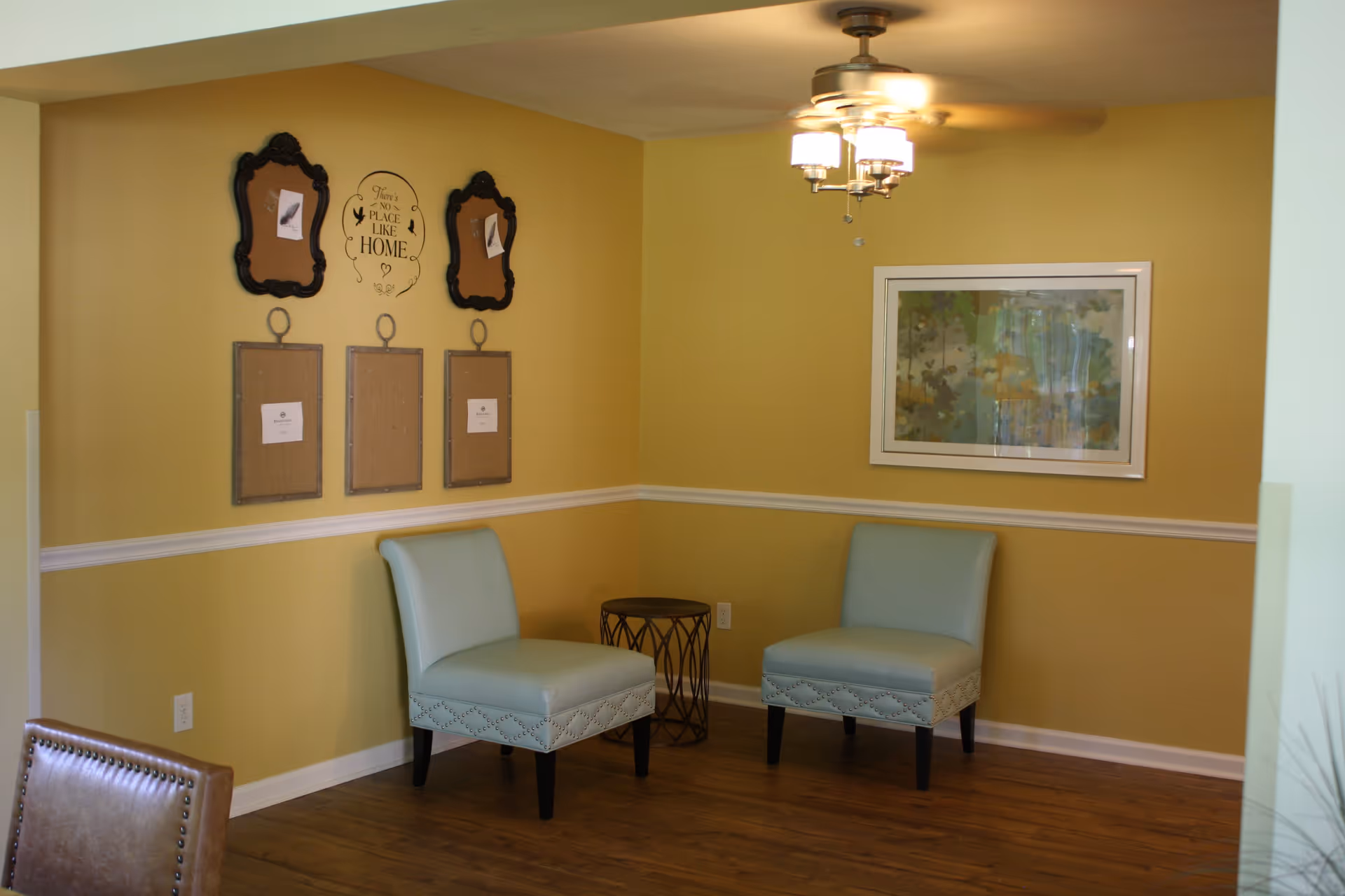 A cozy corner of a room with two light blue upholstered chairs separated by a small round metal side table. The walls are painted yellow with white trim, and there is a ceiling fan with lights above. On the wall, there are decorative frames and a wall decal that reads 'There's no place like home.' A framed abstract painting hangs on the adjacent wall.