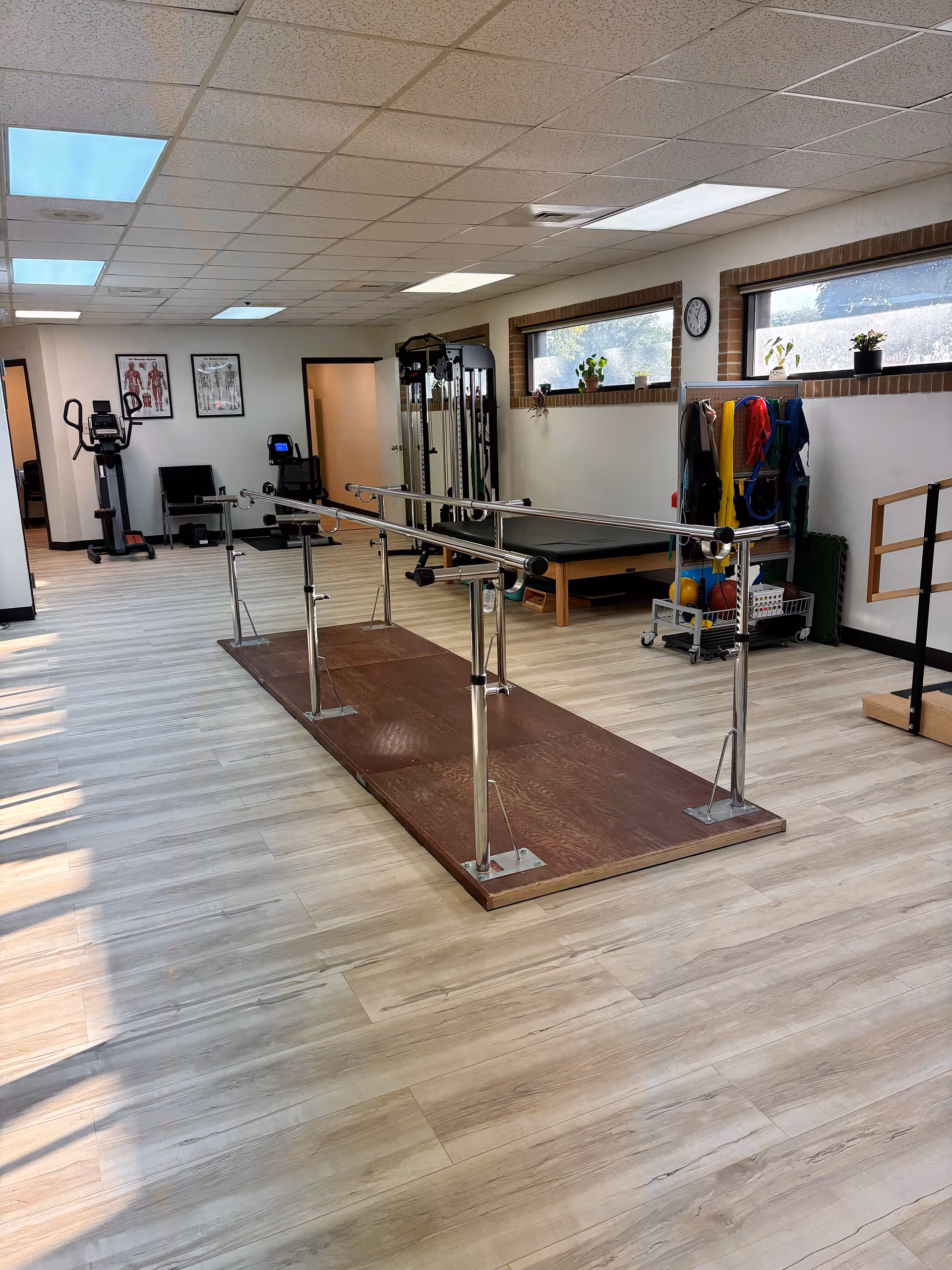 Indoor physical therapy gym with parallel bars, exercise equipment, and large windows.