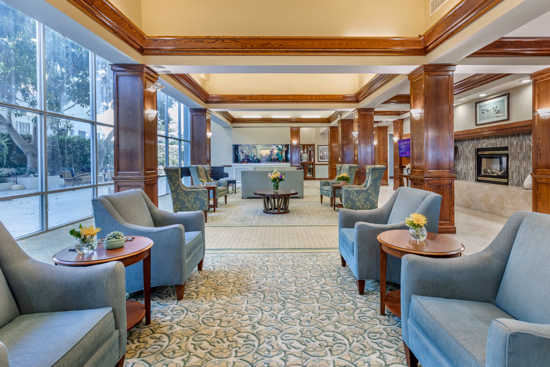 Sunlit communal lounge with blue upholstered chairs, wooden columns, large floor-to-ceiling windows, patterned carpet, and a fireplace.