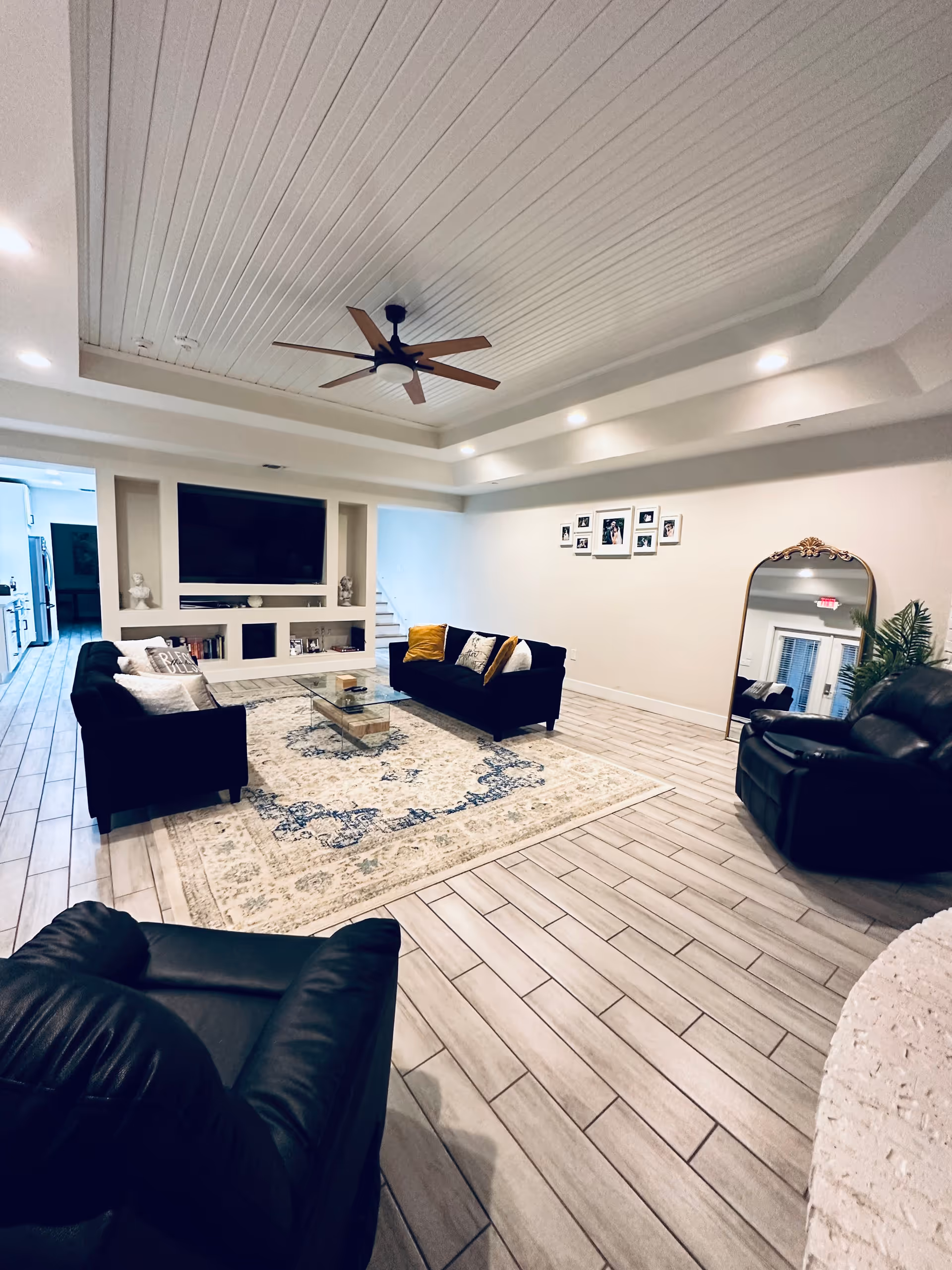 Spacious modern living room with dark sofas arranged around a patterned rug and glass coffee table, built-in TV shelving, ceiling fan, and a large floor mirror.