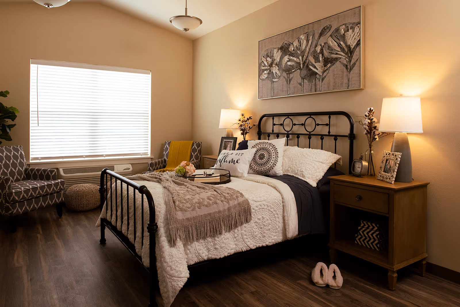 A cozy bedroom with a black metal bed frame, white and beige bedding, decorative pillows, and a beige throw blanket. There are two wooden nightstands with lamps, picture frames, and decorative items. Two patterned armchairs and a round woven ottoman are placed near a large window with blinds. The room has wooden flooring and a large floral painting above the bed.