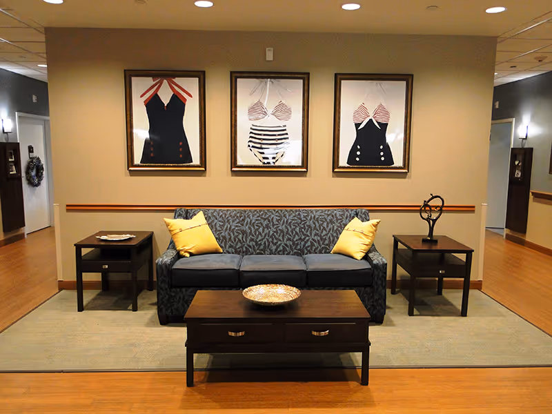 A cozy seating area with a patterned blue sofa adorned with two yellow pillows, flanked by two dark wooden side tables. A dark wooden coffee table with a decorative bowl sits in front of the sofa. Above the sofa, three framed artworks depicting vintage swimsuits hang on a beige wall. The floor is wooden with a large area rug under the furniture, and the space is softly lit with ceiling lights.