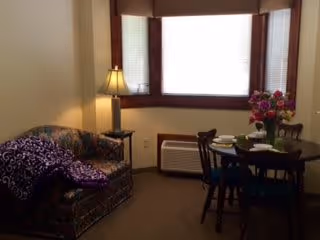 A cozy living area with a floral patterned couch draped with a purple blanket, a small side table with a lamp, and a round dining table set with plates and a vase of flowers near a large window with blinds.