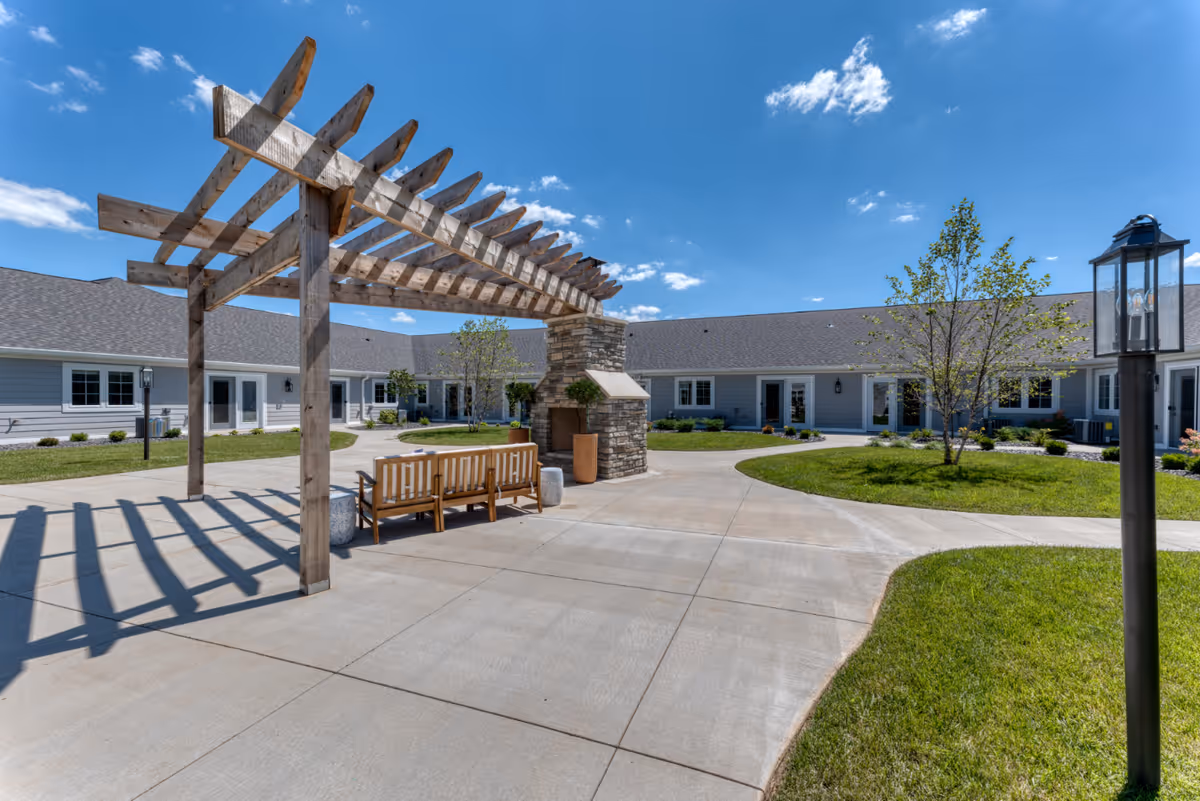 Outdoor courtyard area at Cedarhurst Senior Living of Farmington featuring a wooden pergola with a bench underneath, a stone fireplace, paved walkways, green grass, young trees, and a clear blue sky with scattered clouds.