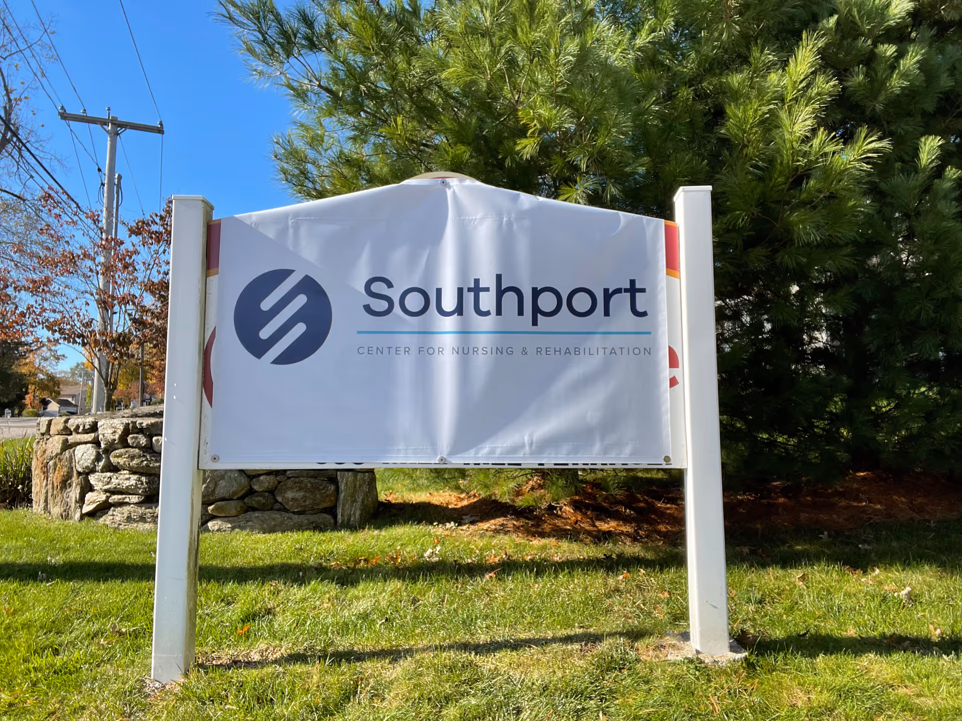 A freestanding outdoor sign reading "Southport Center for Nursing & Rehabilitation" on a grassy lawn with trees and a stone wall behind it.