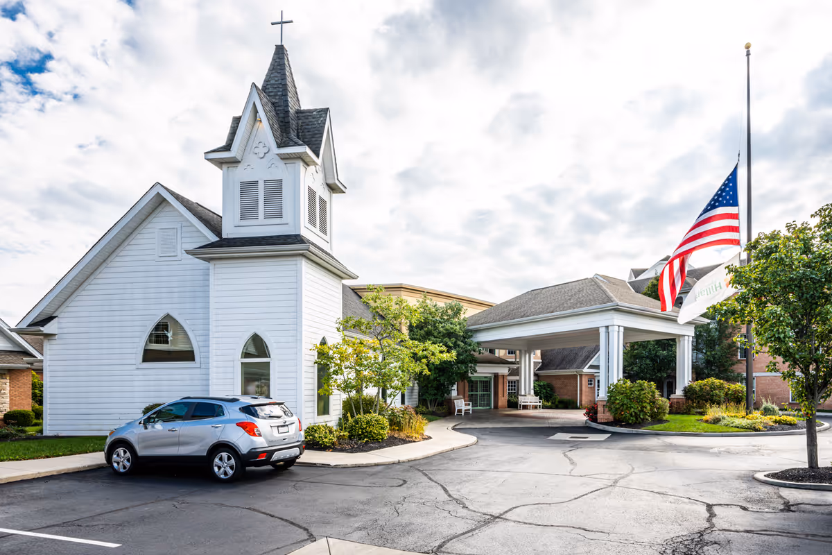 Exterior view of a senior living facility with a white chapel-like building featuring a steeple and cross on the left, a covered entrance with white columns in the center, a silver car parked in front, and an American flag flying at half-mast on the right. The sky is partly cloudy and there are trees and shrubs around the buildings.