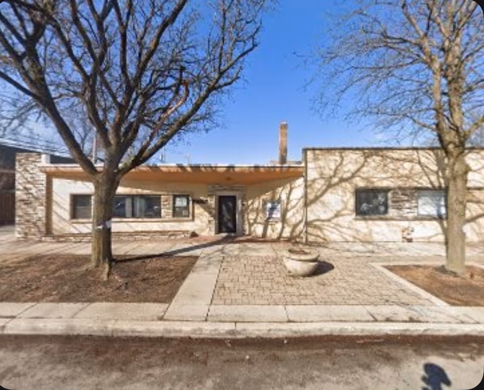 Single-story beige building with a covered entrance and leafless trees in a paved front yard.