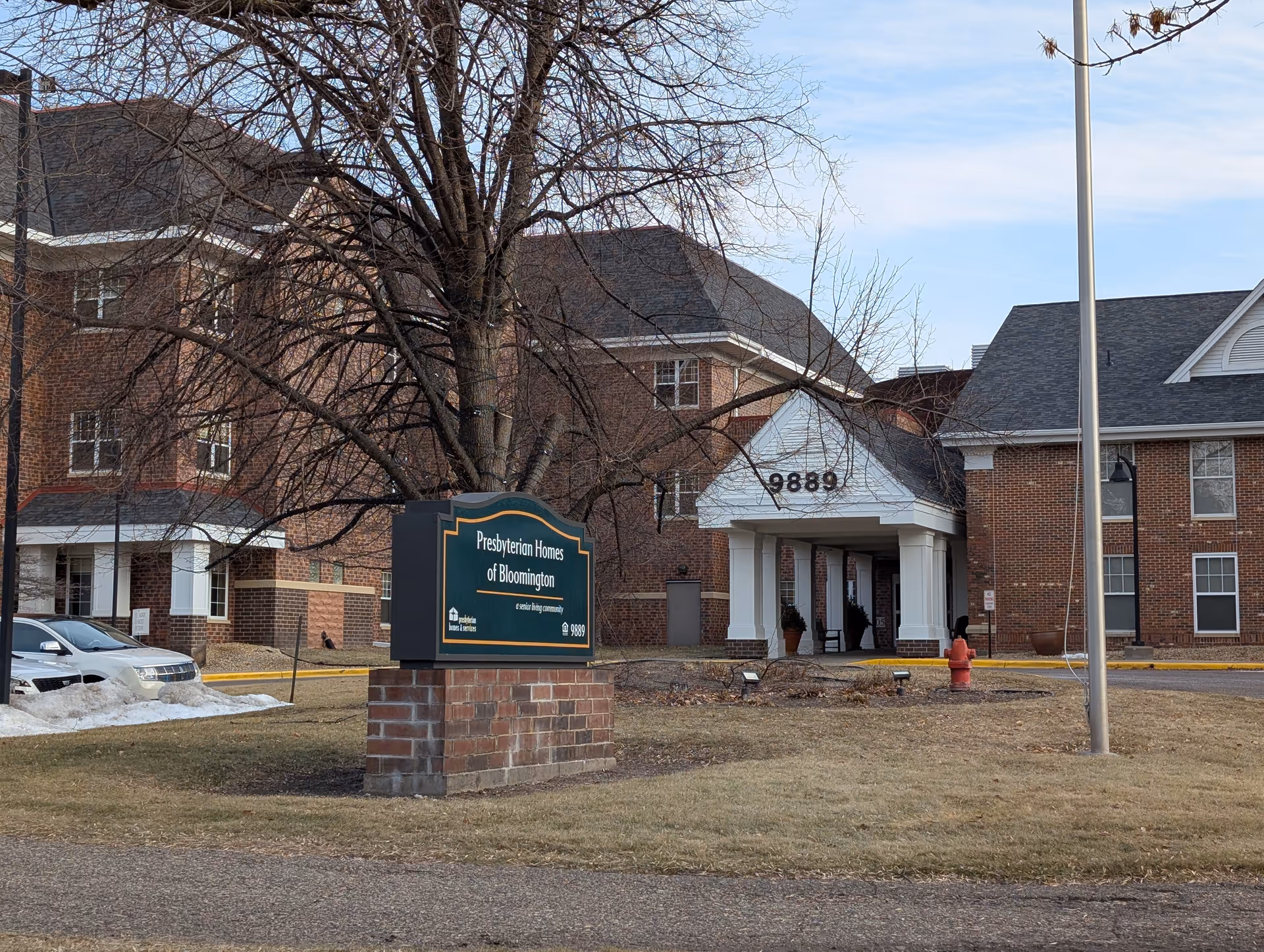 Front exterior of Presbyterian Homes of Bloomington showing a brick sign, the building entrance with a gabled portico, and a flagpole.