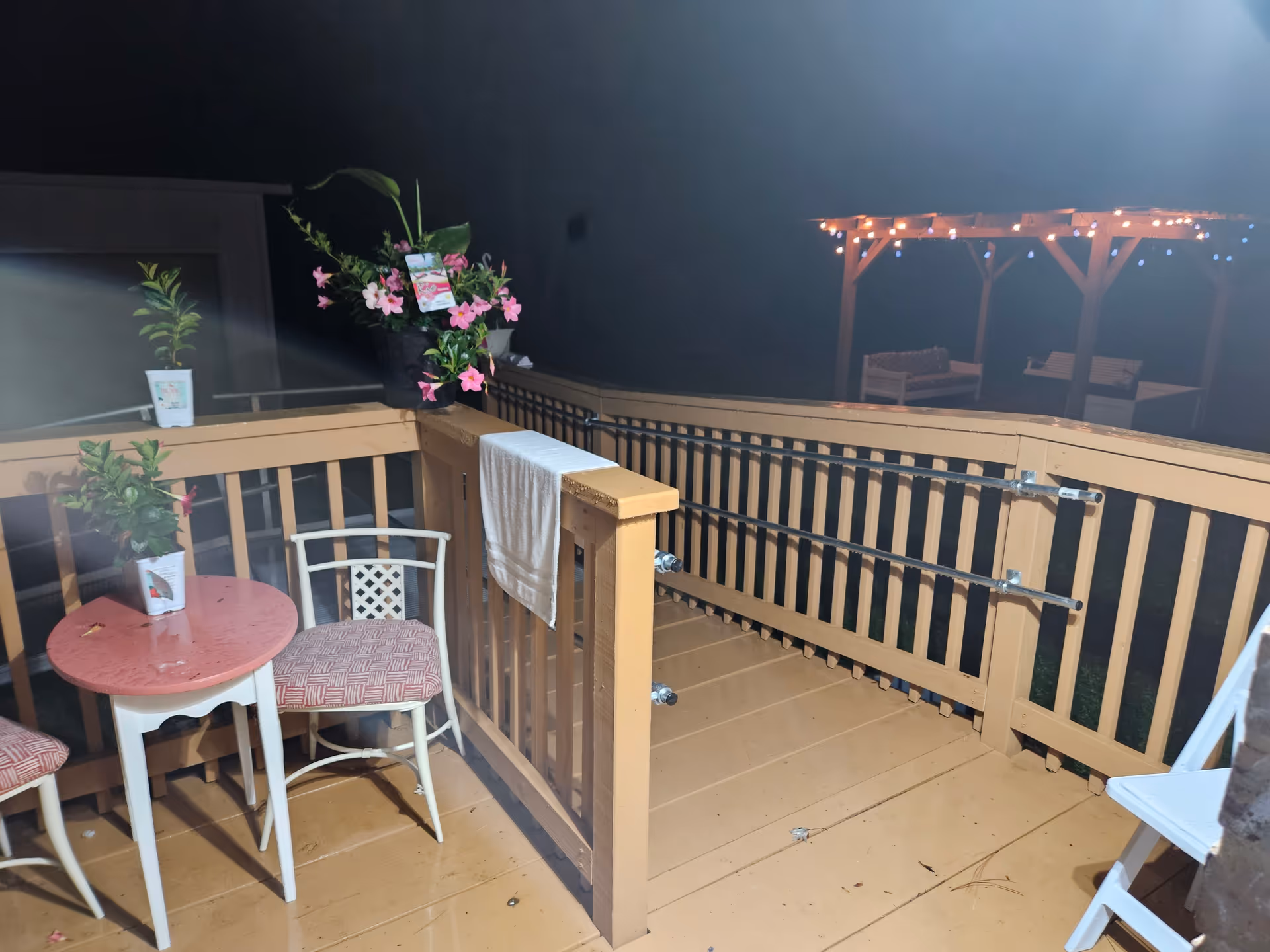 Night outdoor balcony with a small round table, two chairs, plants, and string lights.