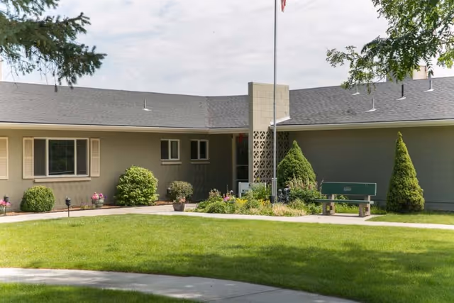 Front of a single-story care facility with a green lawn, flagpole, bench, shrubs, and an entrance alcove.