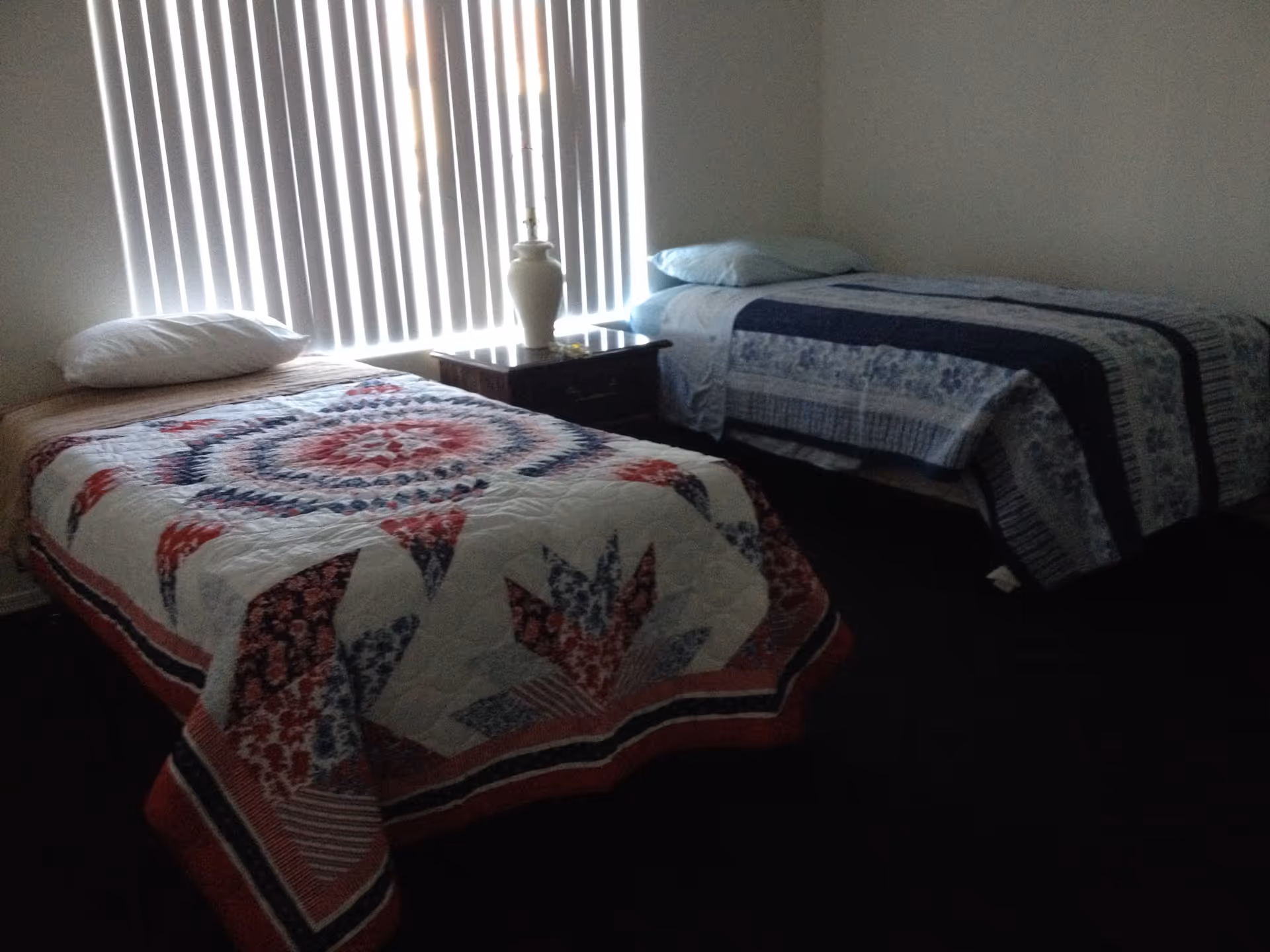 A bedroom with two single beds, each covered with patterned quilts. A small wooden nightstand with a white lamp is positioned between the beds. Vertical blinds cover the window behind the beds.