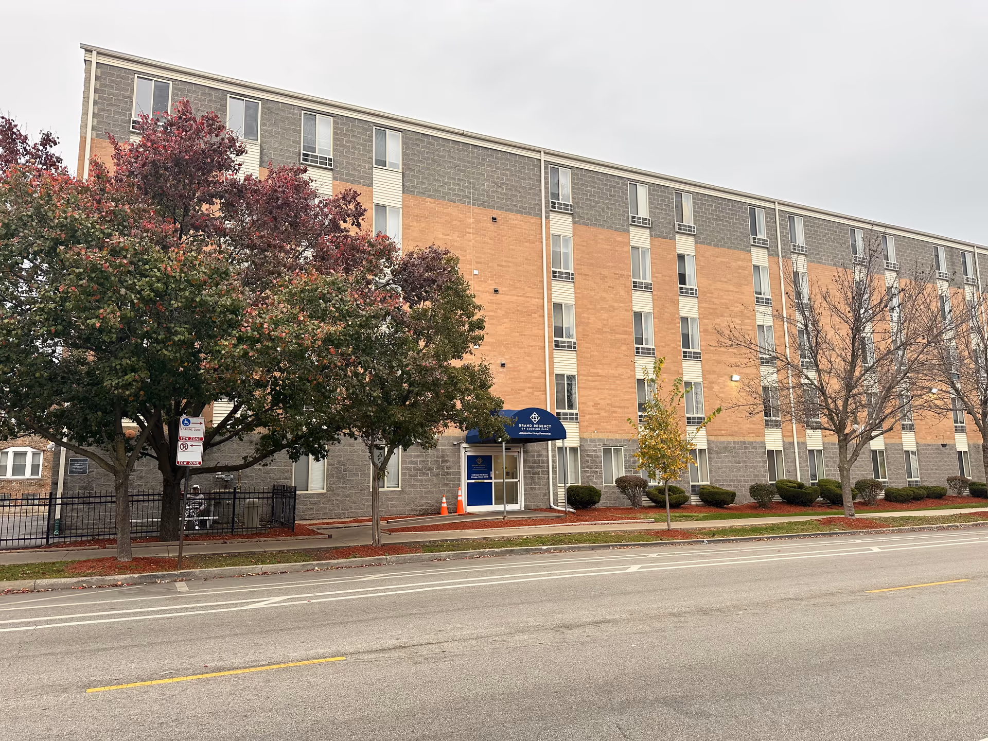 Exterior view of a multi-story brick and concrete building with several windows. The entrance has a blue awning with the text 'Grand Regency of Jackson Park Supportive Living' and some orange traffic cones nearby. Trees with autumn foliage line the sidewalk in front of the building, and a street runs parallel to the building.