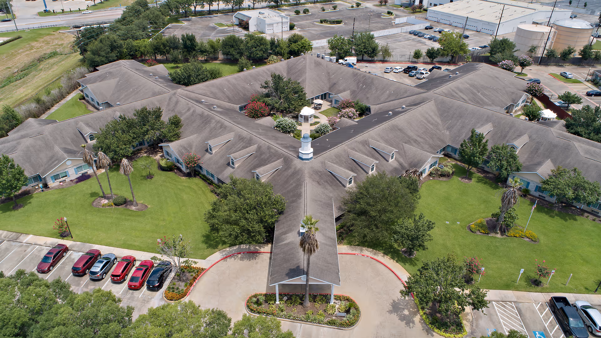 Aerial view of Morada Cy-Fair senior living facility showing a large, single-story building with a Y-shaped layout surrounded by green lawns, trees, and landscaped areas. Several cars are parked in the parking lot adjacent to the building, and there are paved walkways and driveways leading to the entrance.