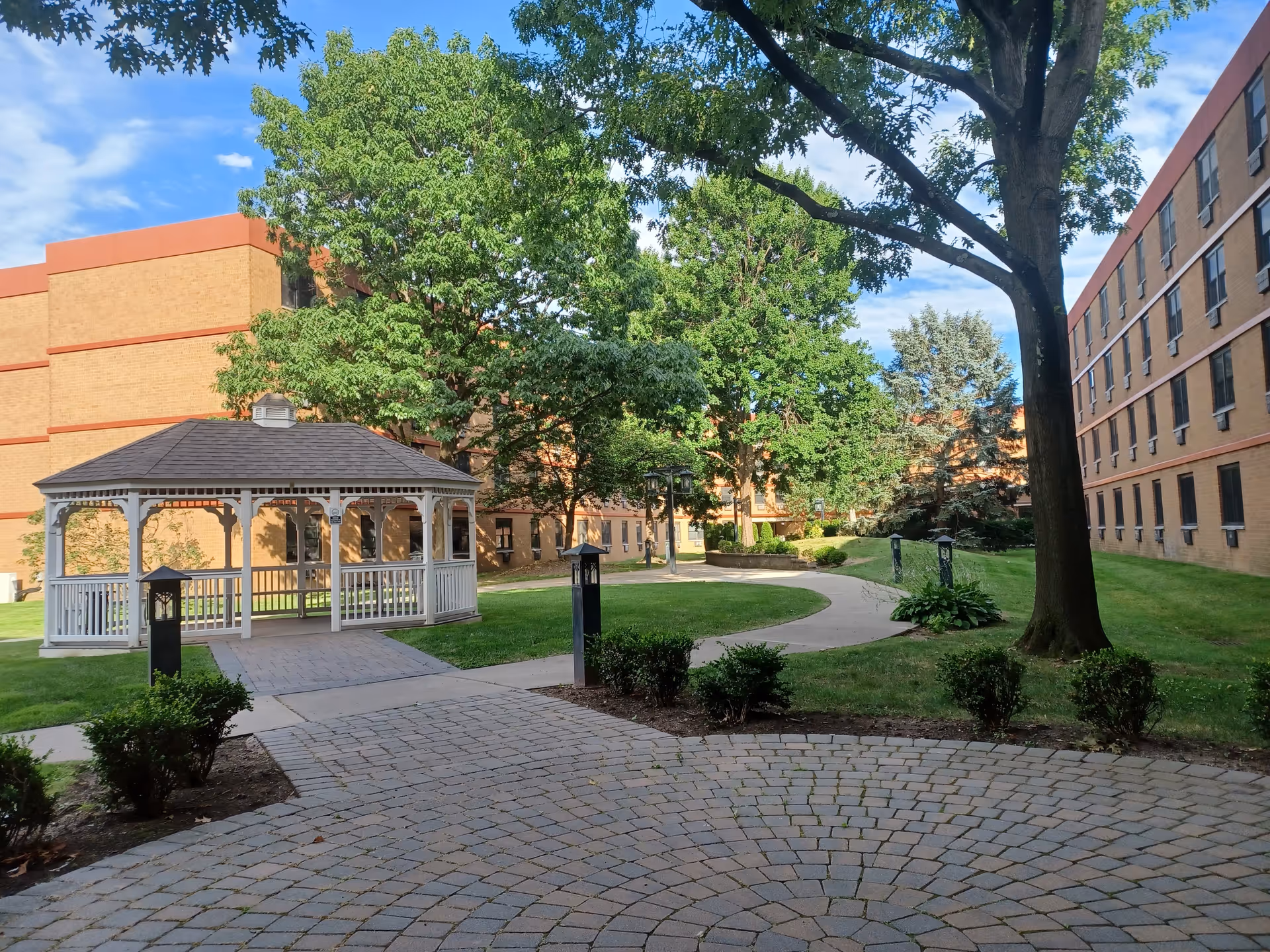 A serene senior living courtyard with a white gazebo, brick buildings, trees, and a winding path.