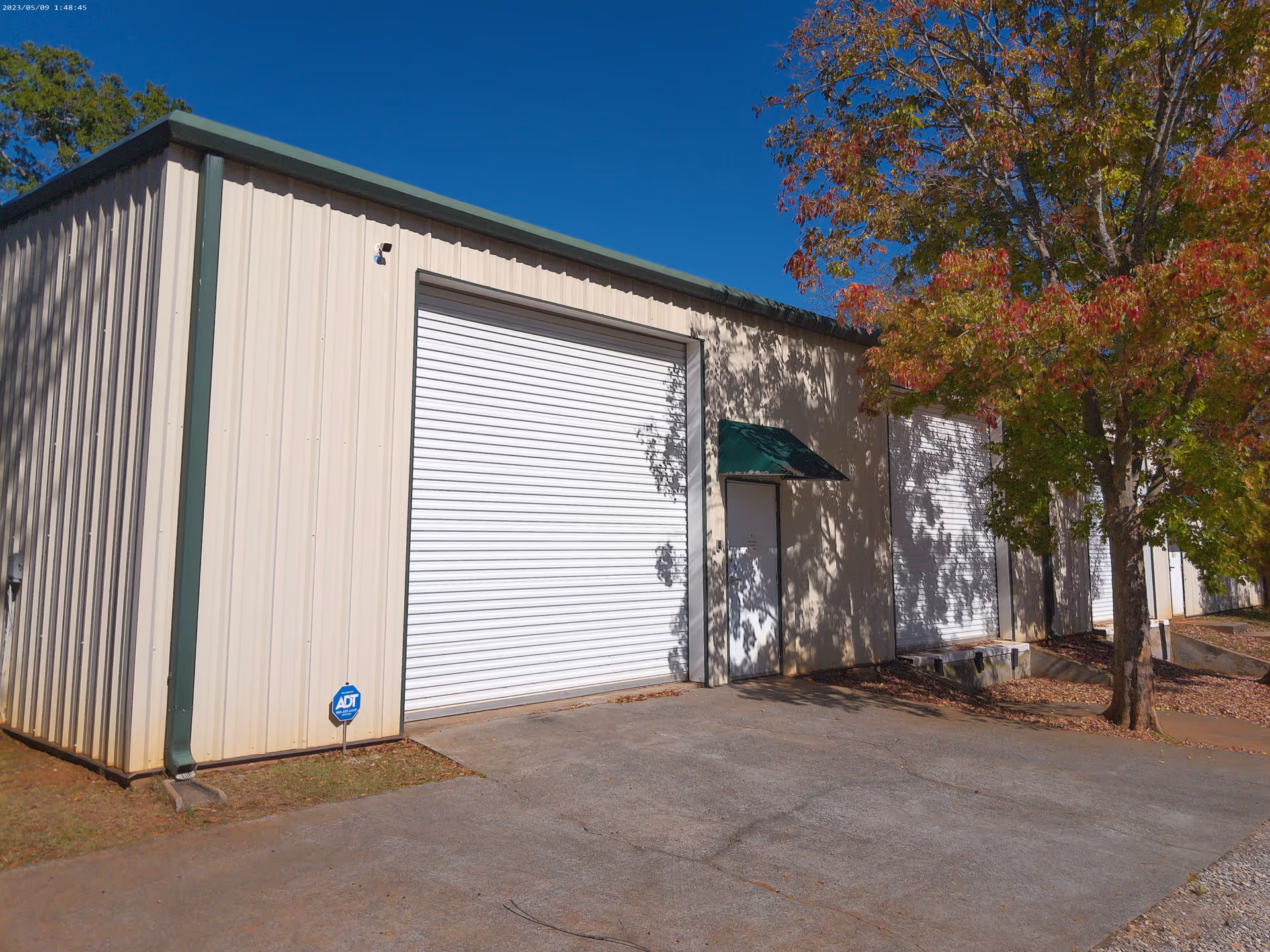 Exterior view of a beige metal building with large white rolling doors and a tree.