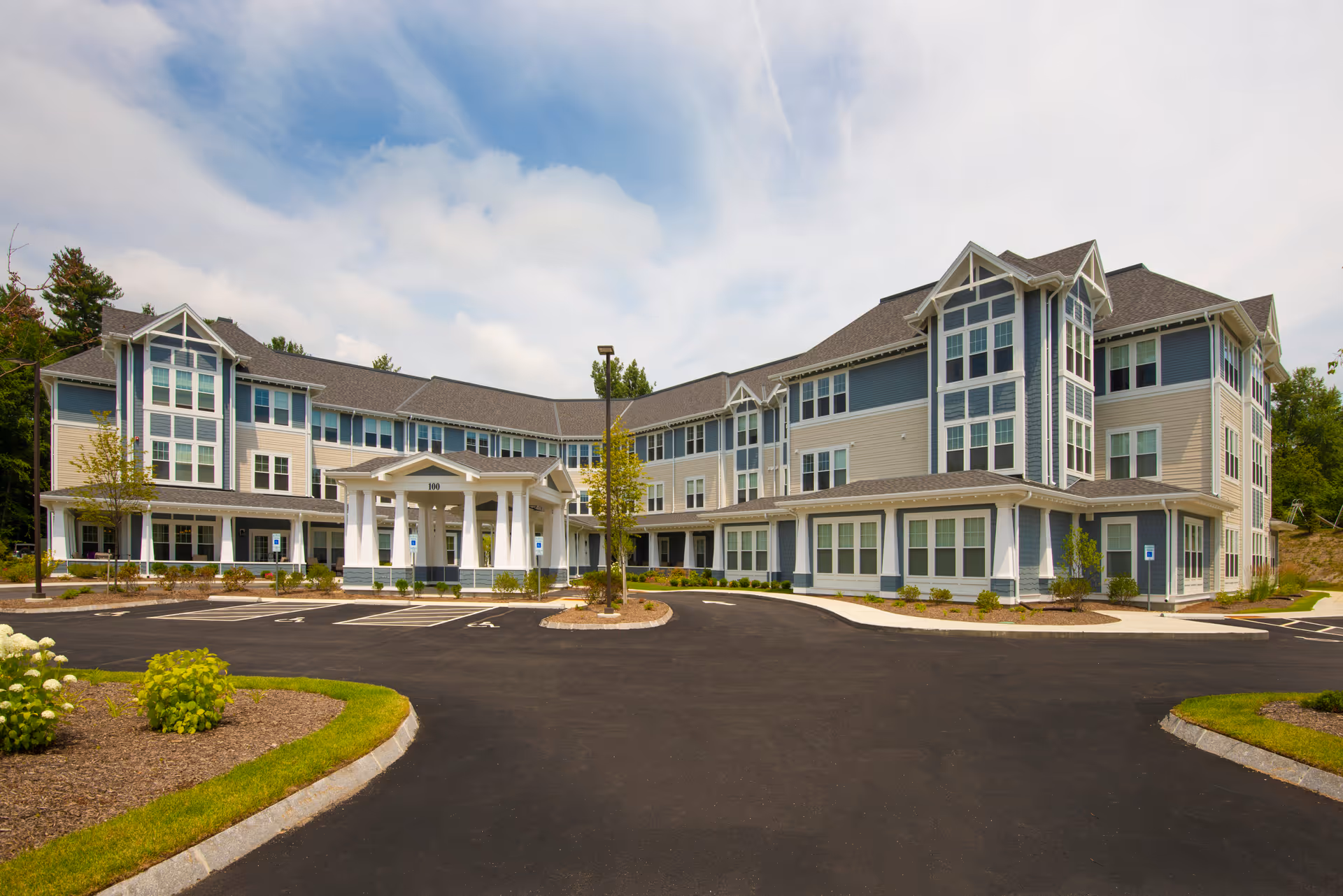 Three-story senior living building with a covered porte-cochere entrance, landscaped grounds, and a paved parking area under a partly cloudy sky.
