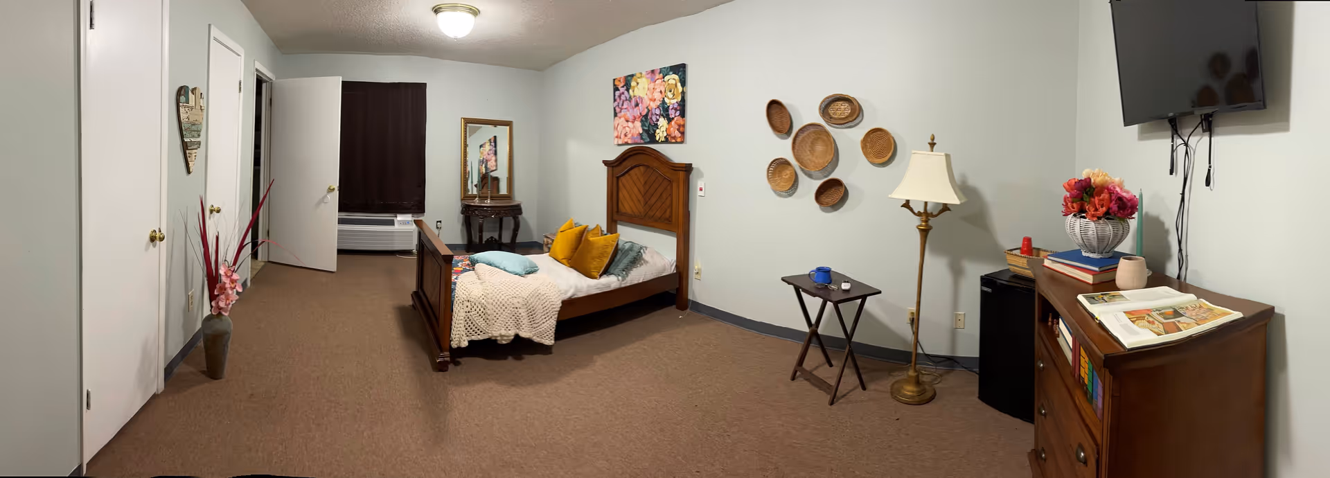 A cozy bedroom featuring a wooden bed with colorful pillows, a decorative mirror, and a wall adorned with woven baskets. The room includes a small table with a blue cup, a lamp, and a dresser with books and flowers.