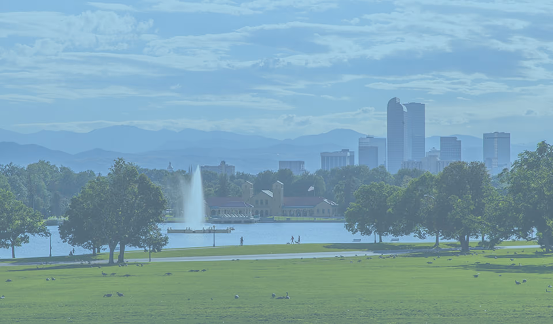 Lush park with a lake, fountain, trees, and distant city skyline.