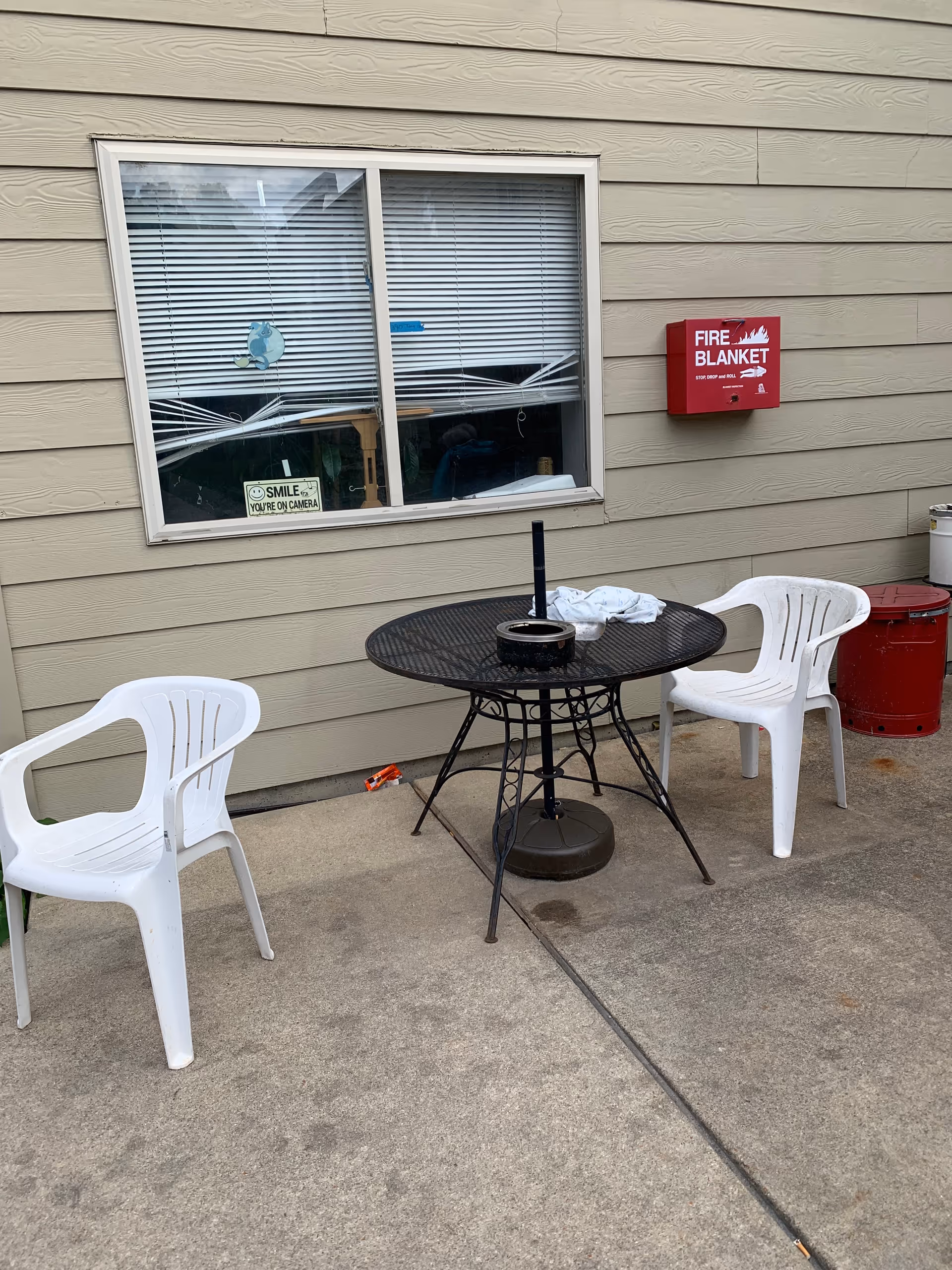 Outdoor patio with a round black table and two white plastic chairs beside a beige building.