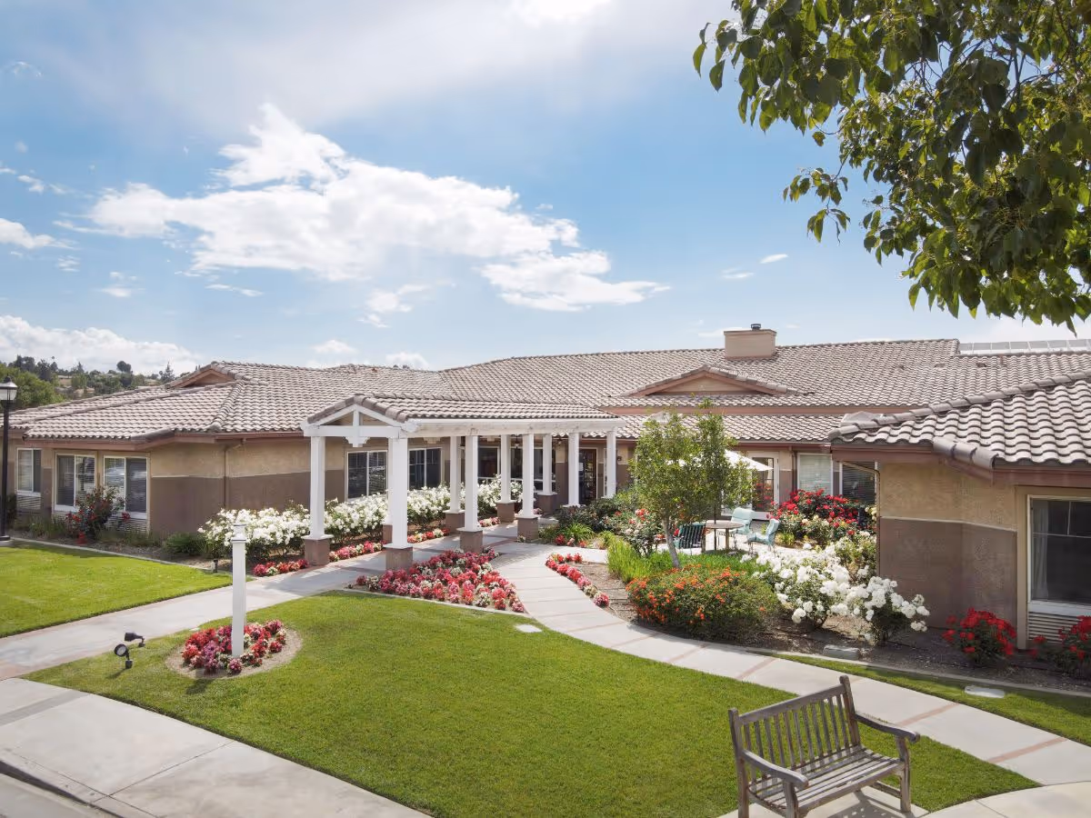 Front entrance of a single-story senior living building with a covered walkway, well-kept lawns and flowerbeds, and a bench under a sunny sky.