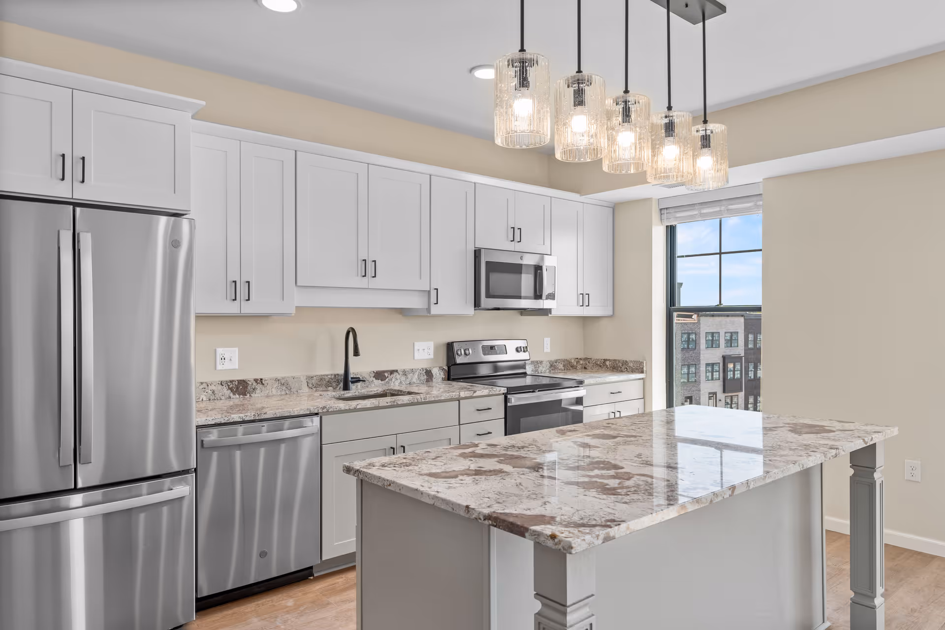 Modern kitchen with white cabinets, stainless steel refrigerator, dishwasher, oven, and microwave. A large kitchen island with a marble countertop is in the center, illuminated by five pendant lights. A window shows a view of nearby buildings.